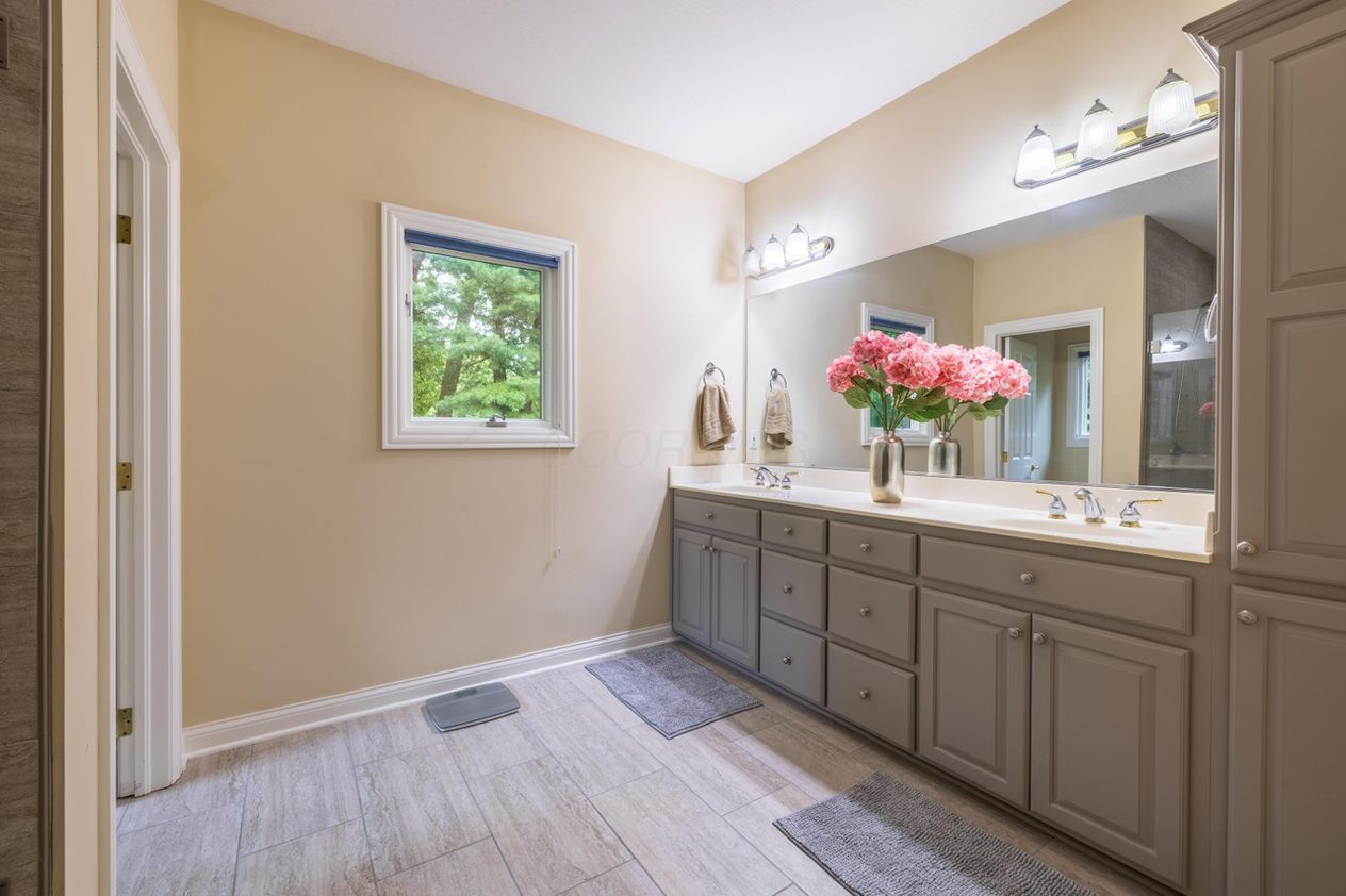 The Simmons master bath before work — cream walls, a grey raised-panel double vanity with a white cultured-marble top, a bulb-strip light bar over a plain plate-glass mirror, and a white-framed window.