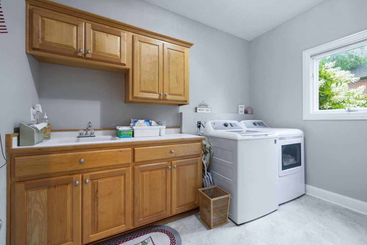 The Simmons laundry room before work — honey-oak raised-panel cabinets, a white countertop with a chrome faucet, bare painted wall with no backsplash, and a white washer and dryer.