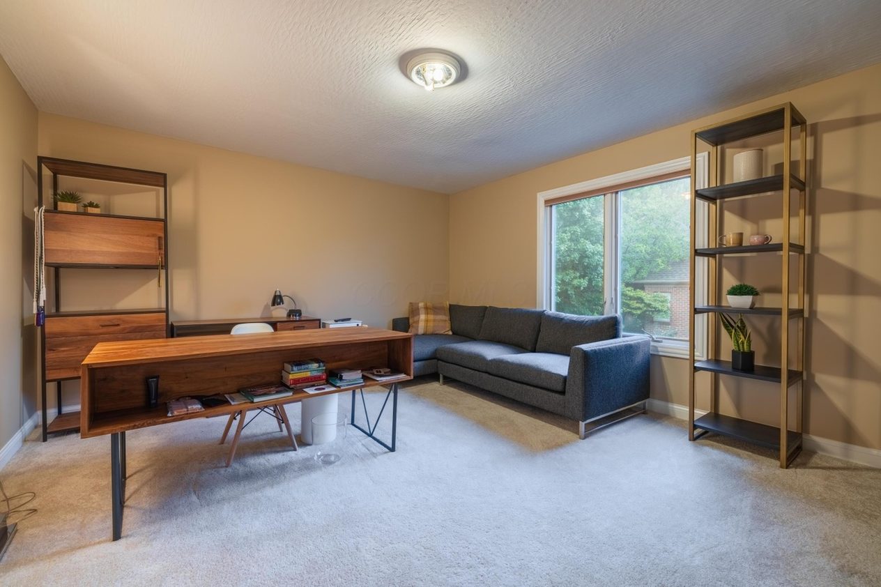 The Simmons home office before work — beige walls, pale wall-to-wall carpet, freestanding shelf units and a wood desk, and a small flush-mount ceiling light.